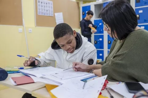 Un jeune est en train de faire ses devoirs, accompagnée d'une jeune femme dans une salle de classe